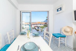 Un comedor con una mesa dispuesta para dos personas, vista al mar y edificios, y una silla con cojín azul.