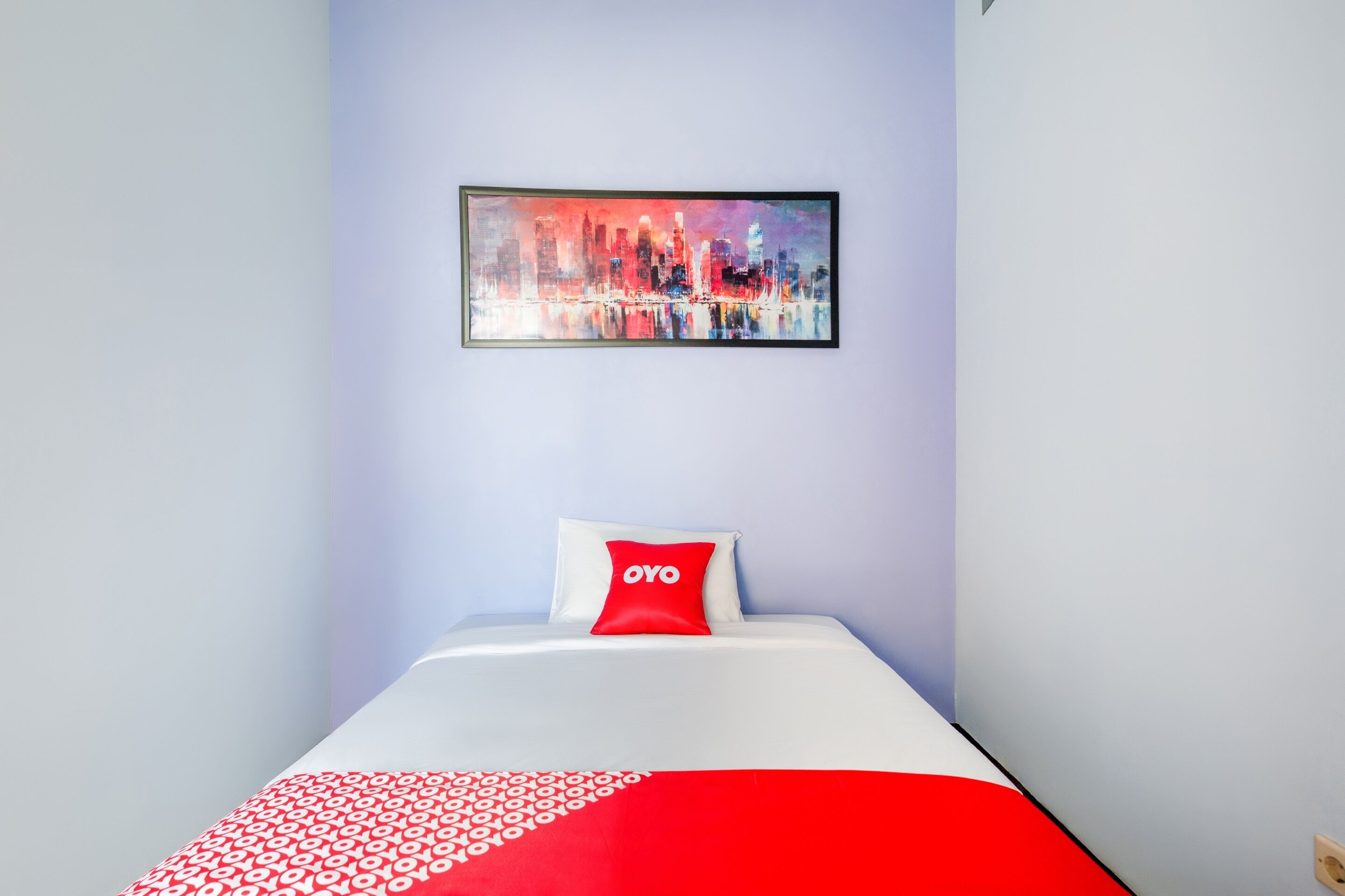 Ein Hotelzimmer mit einem Bett, einer rot-weißen Bettdecke, einem roten Kopfkissen mit dem Schriftzug "OYO" und einem Stadtbild-Gemälde an der Wand.