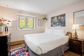 A bedroom with a large bed, a window with a view of greenery, a painting on the wall, and a bedside table with a lamp. - Snowmass Mountain D1 (Snowmass Village)
