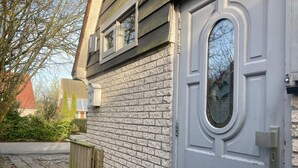 Un edificio residencial con una puerta gris, una ventana con marco decorativo y un muro de ladrillo.