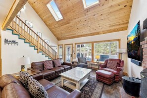 A spacious living room with a high ceiling, wooden beams, a staircase, and a large window offering a view of the outdoors. - 6BR Tahoe Cabin, Hot Tub, Game Room, Near Heavenly (South Lake Tahoe)