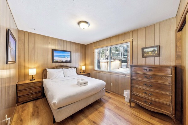 A hotel room with a wooden paneled wall, a bed with white linens, a wooden dresser, a nightstand with a lamp, and a window with blinds.