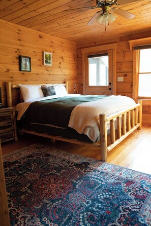Une chambre dans une cabane en bois, avec un lit, une table de chevet, une fenêtre et un ventilateur de plafond.