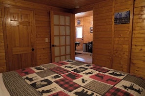 A wooden cabin interior with a quilted bedspread, a door leading to another room, and a framed picture on the wall.