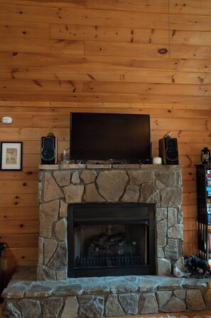 A stone fireplace with a flat-screen TV on top, a stone mantle, and a framed picture to the left. - Babblebrook Lodge Creekfront Cabin w/River Access Close to Town (Murphy)