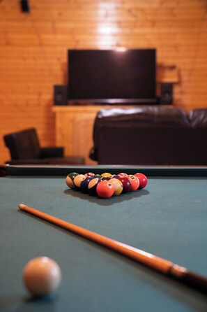 A pool table with balls and a cue stick, a television mounted on the wall, and a leather sofa in a room with wooden paneling.