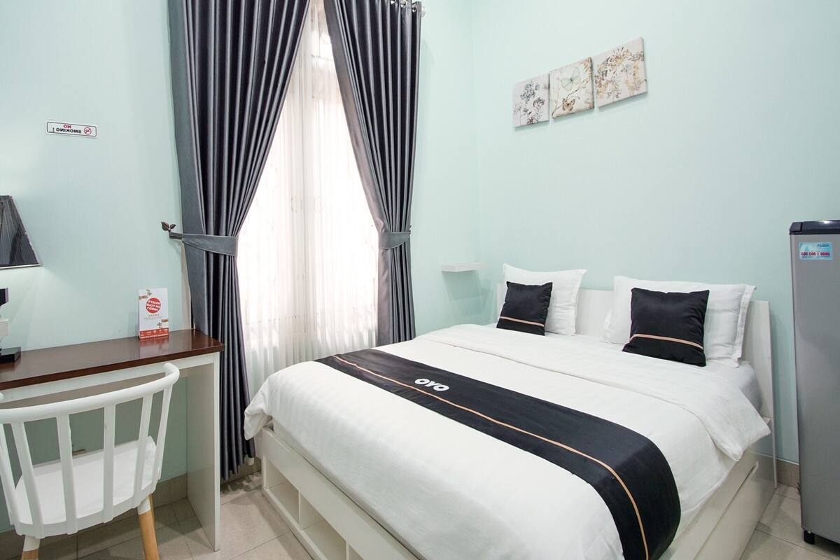 Hotelkamer met een bed, een bureau en een stoel. Gordijnen hangen voor het raam, en een koelkast is aan de rechterkant van de kamer te zien.