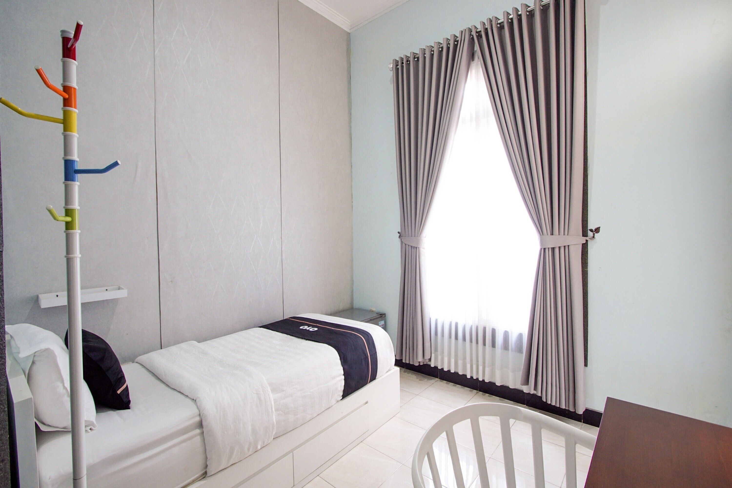A small, neatly arranged bedroom with a single bed, a bedside table, a chair, and a window with curtains.