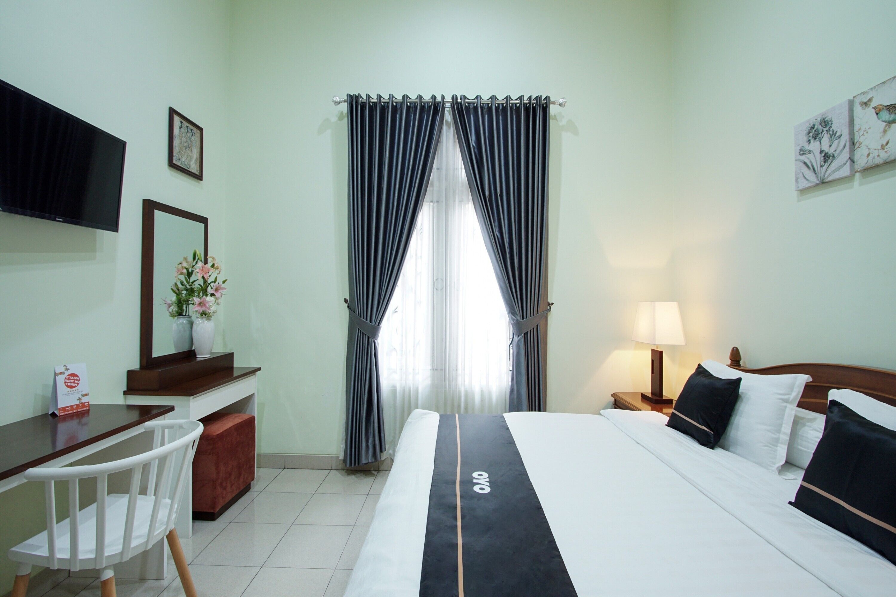 A hotel room with a bed, a desk, a chair, a TV, a window with curtains, and a nightstand with a lamp.