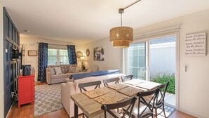 A cozy living room with a sofa, a dining table with chairs, a hanging light fixture, and a sliding glass door leading to a garden. - River Shores Cottage (Ottawa)