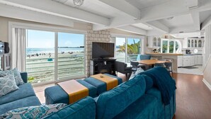 A spacious living area with a large window overlooking a beach, a cozy seating area, and a kitchen in the background. - Rio Del Mar Boathouse - steps from the beach (Aptos)