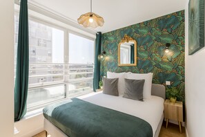 A bedroom with a large bed, a window with curtains, a wall with a leafy pattern, a mirror, and a small bedside table with a plant.