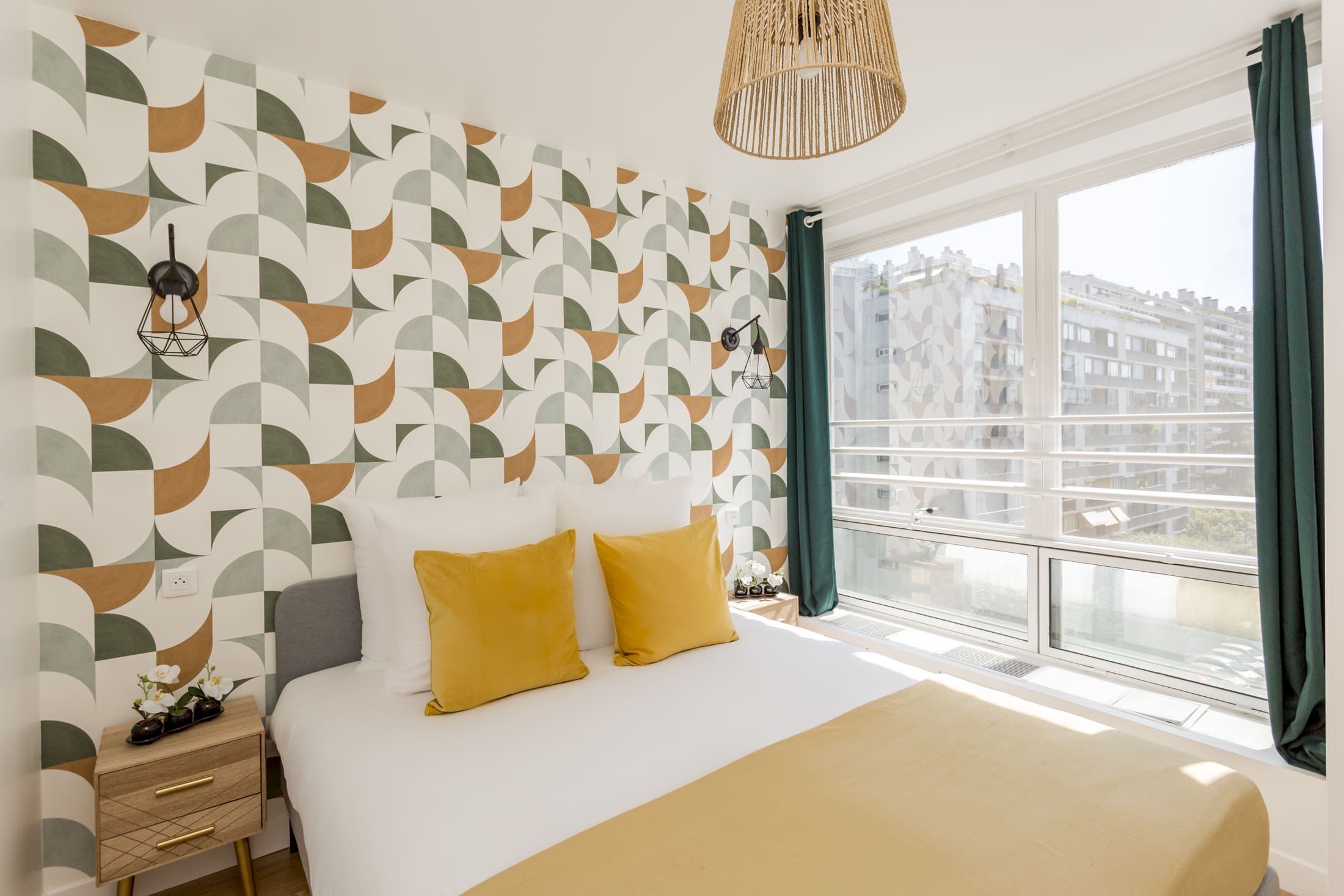 A hotel room with a geometric patterned wall, a bed with two yellow pillows, a bedside table with a lamp, and a view of buildings through a window.
