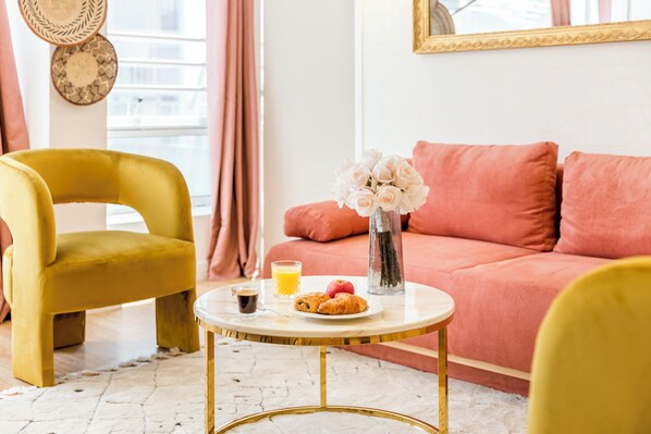 Une pièce chaleureuse avec un fauteuil jaune, un canapé rose et une petite table ronde sur laquelle sont disposés des éléments pour le petit-déjeuner.