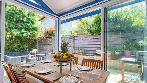 A dining area with a wooden table set for four, surrounded by wicker chairs. A fruit basket is placed on the table, and the room has large windows offering a view of the garden and trees outside.