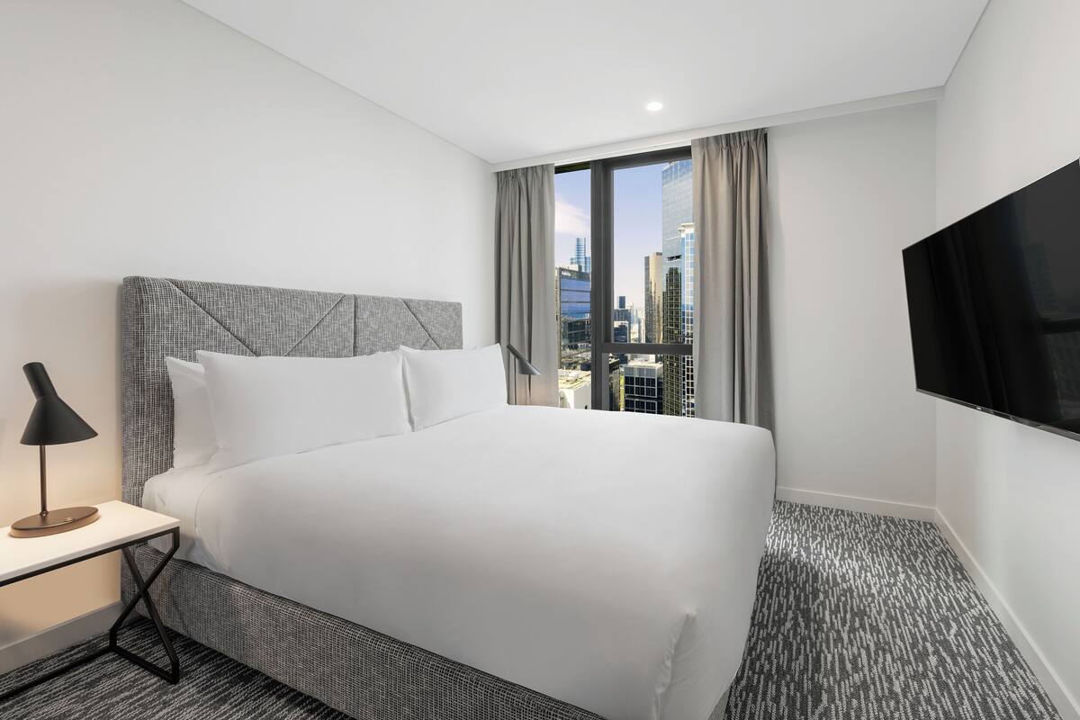 A modern hotel room with a large bed, a bedside table with a lamp, a flat-screen TV, and a city view through a large window.