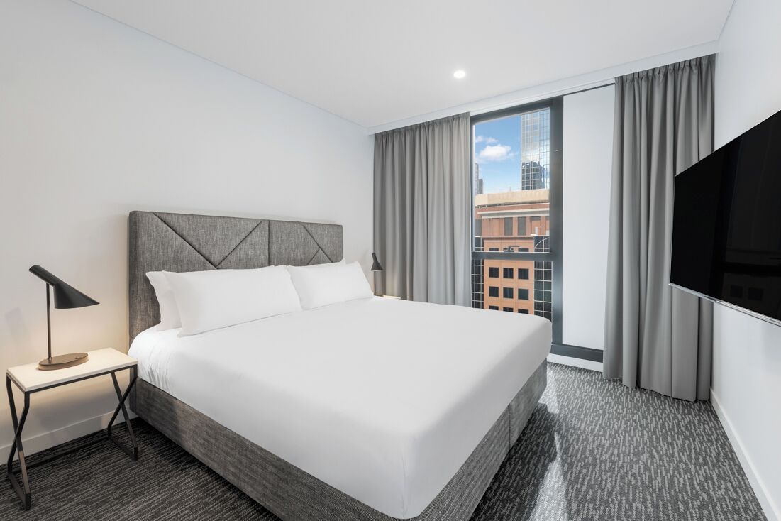 A hotel room with a large bed, a TV, a nightstand, and a view of the city.