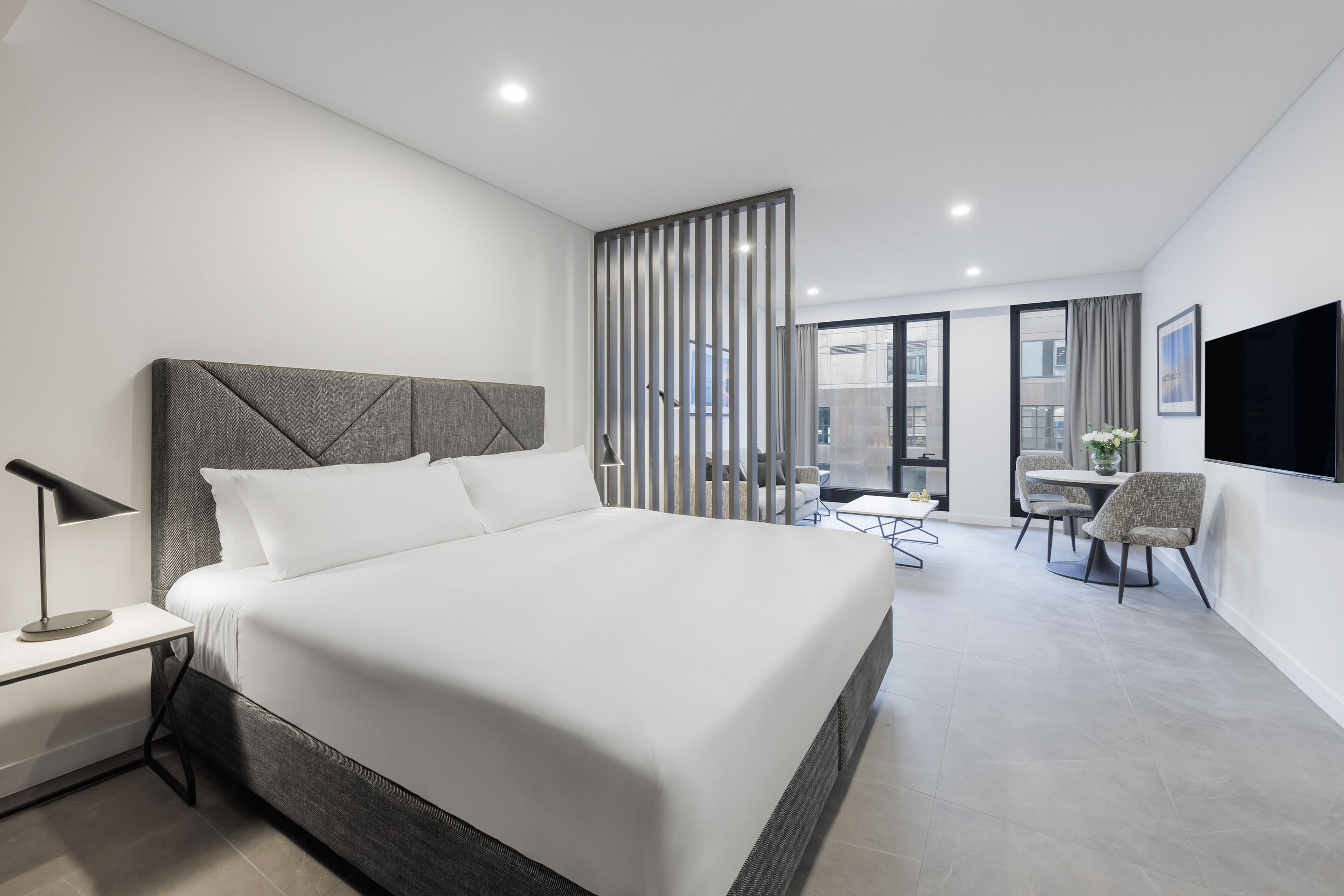 Une chambre d’hôtel moderne dotée d’un grand lit, d’un bureau avec une lampe, d’un coin repas avec des chaises et d’un téléviseur fixé au mur.