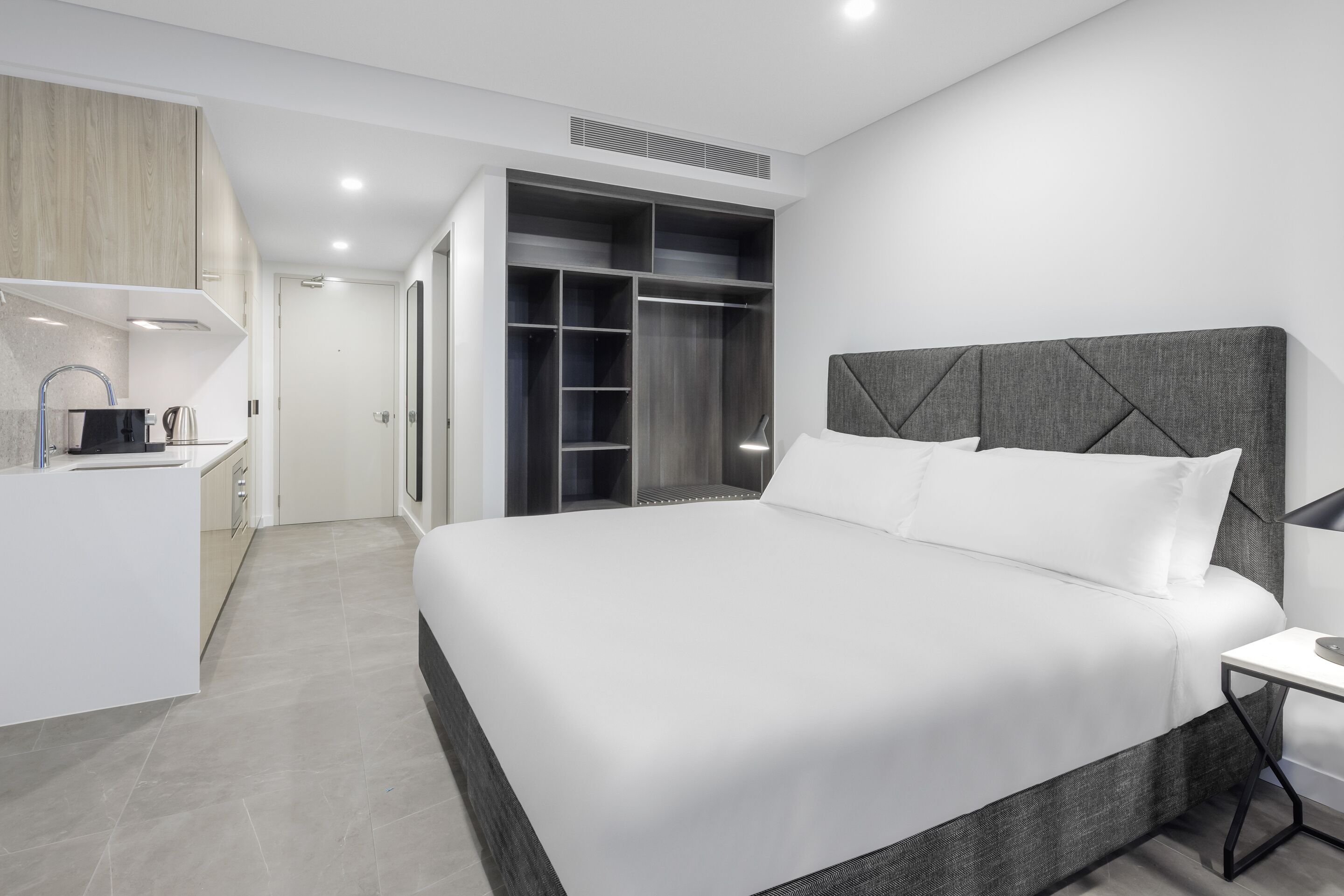A modern hotel room with a large bed, a built-in wardrobe, a small kitchenette, and a bedside table.