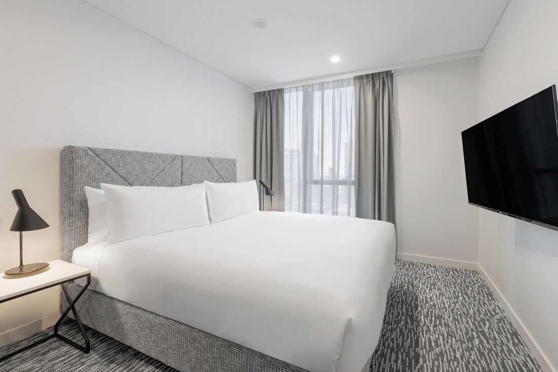 A hotel room with a large bed, a flat-screen TV mounted on the wall, a bedside table with a lamp, and a window with curtains.
