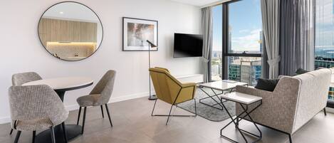 A modern hotel room with a dining area, a sofa, a TV, and a large window with a city view.
