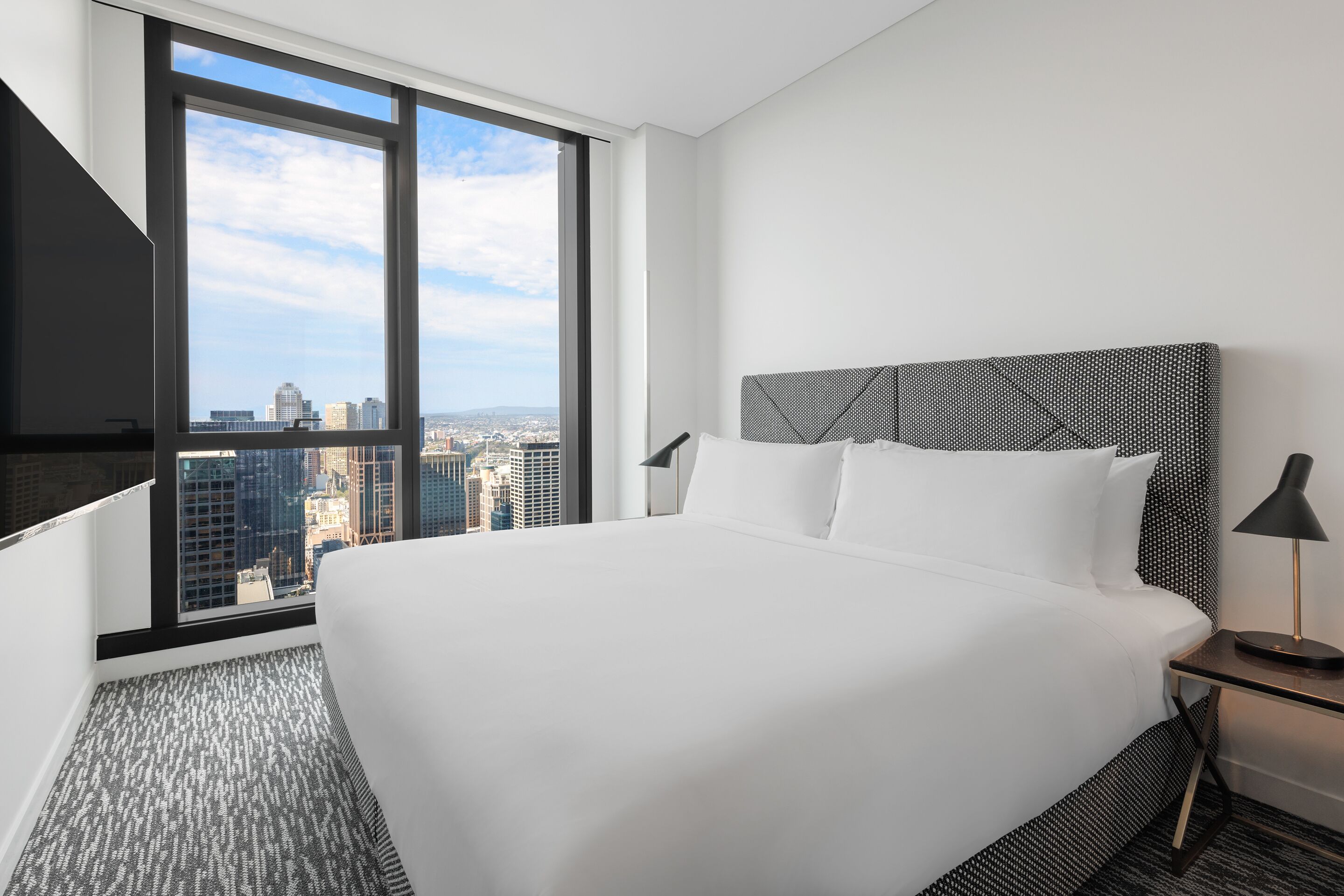 A modern hotel room with a large bed, a TV mounted on the wall, and a city view through large windows.