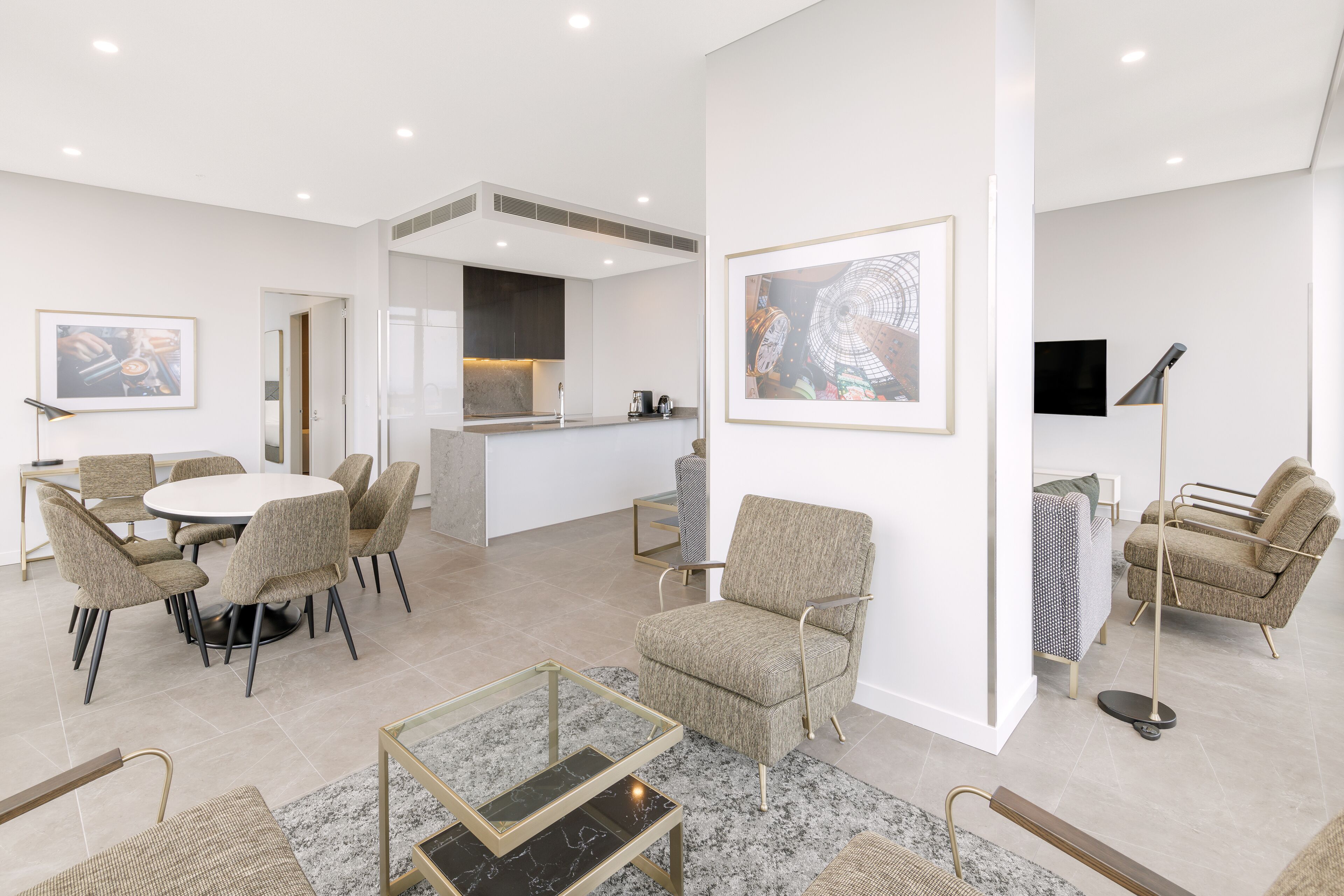 A modern living room with a round dining table, comfortable seating, a kitchen area, and artwork on the walls.