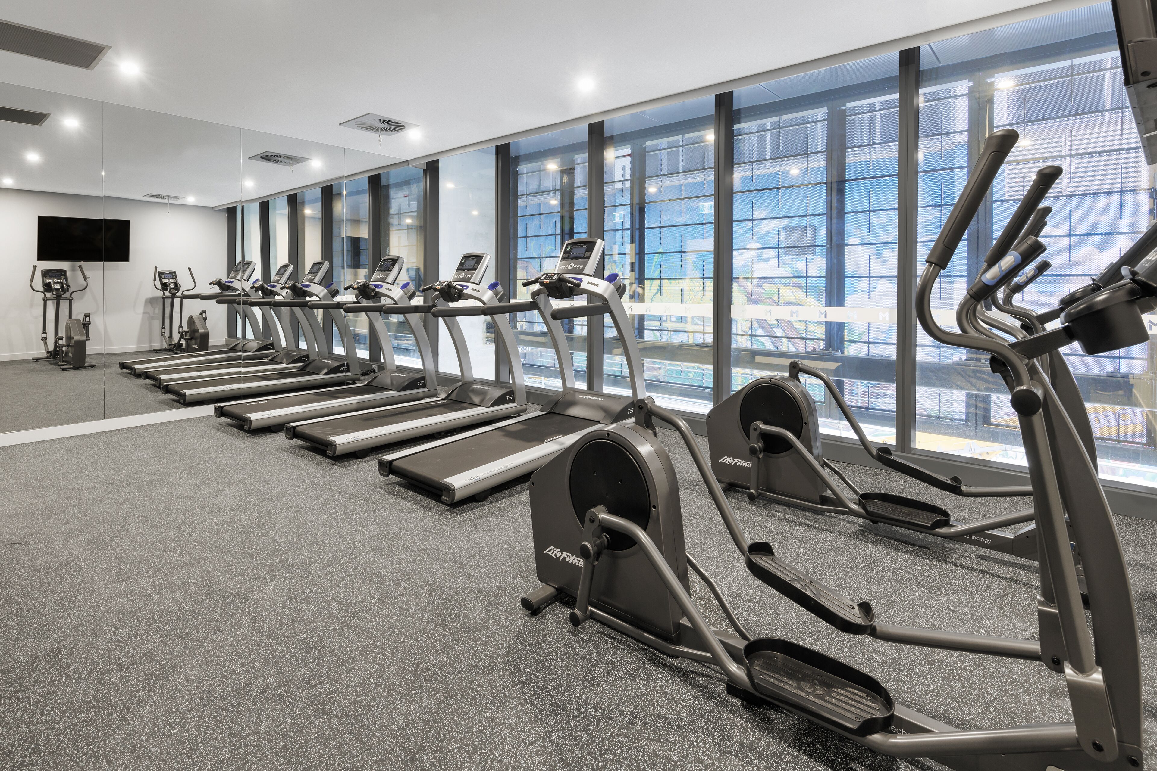 Un gimnasio con cintas de correr, elípticas y una bicicleta estática, ventanas amplias y una televisión de pantalla plana.