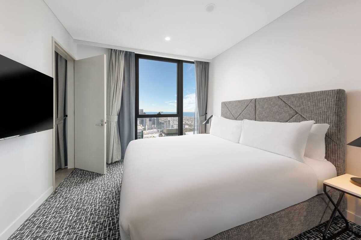 A hotel room with a large bed, a flat-screen TV, and a city view through a large window.