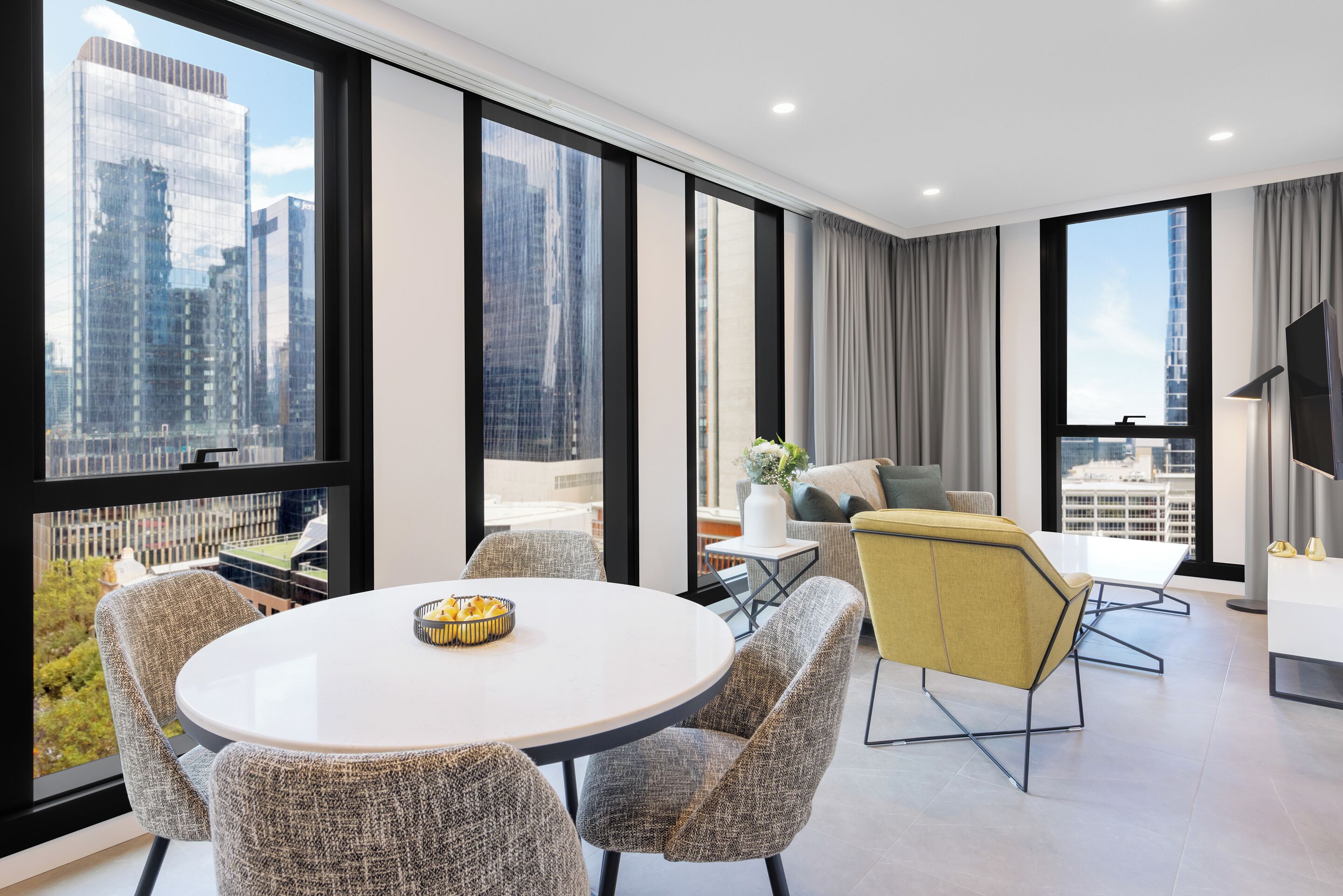 A modern hotel room with a round dining table, grey upholstered chairs, a yellow accent chair, a sofa, and a TV on a stand.