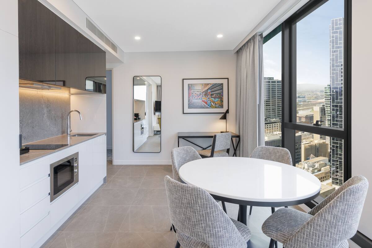 A modern kitchen with a microwave, a dining area with a round table and four chairs, a large mirror, and a city view through a large window.