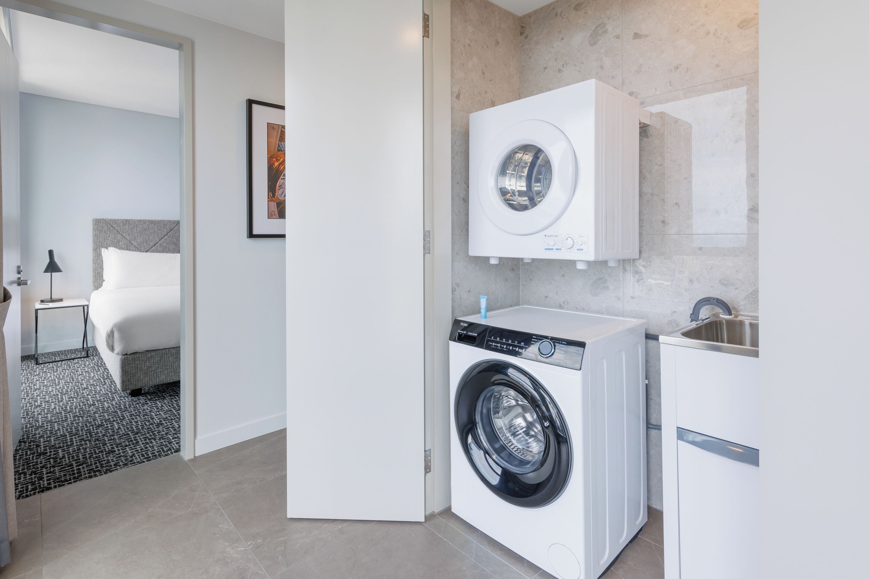 A modern hotel room with a washing machine, a bed, a small table with a lamp, and a framed picture on the wall.