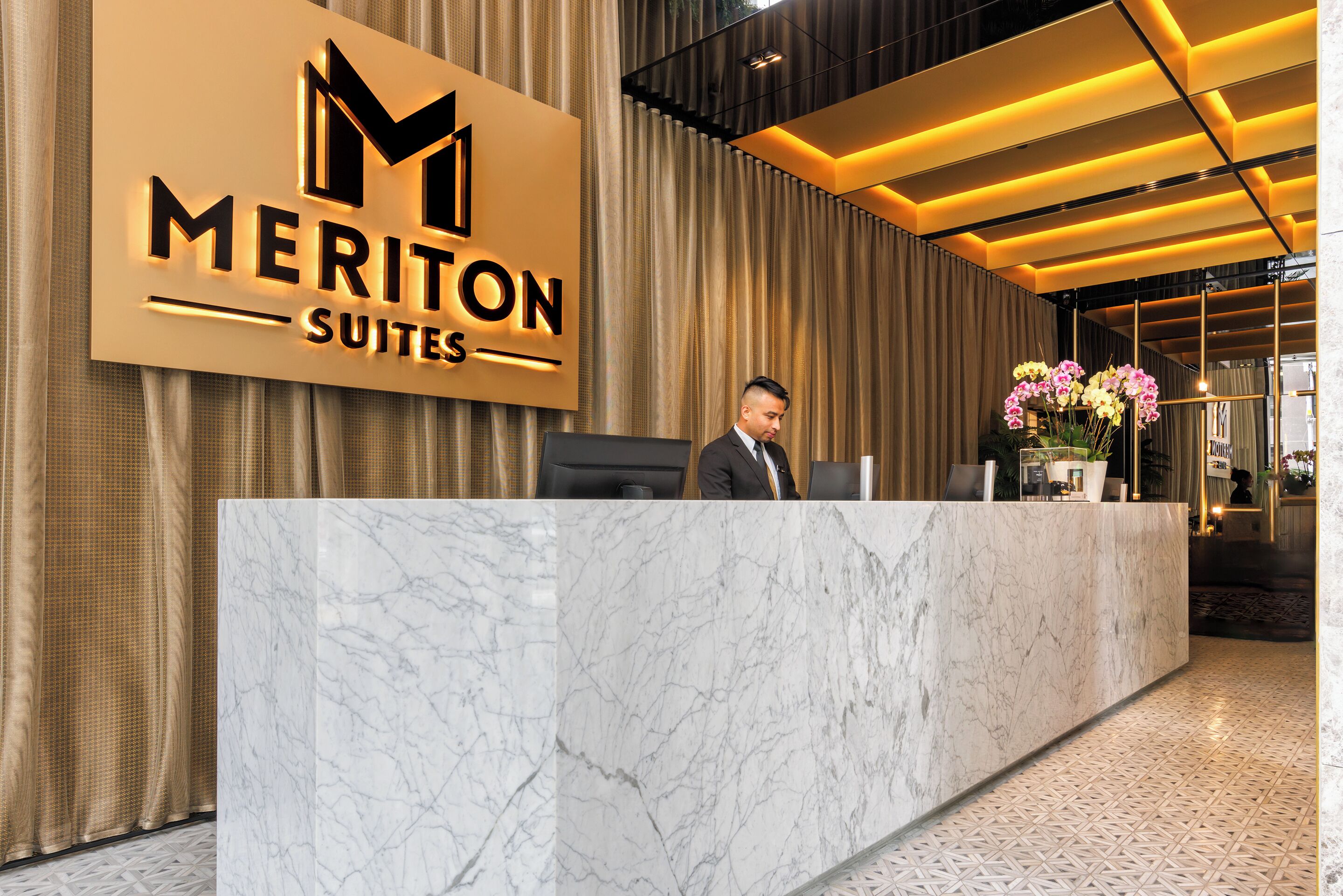 A reception desk with a marble top, a large sign for Meriton Suites, and a modern interior design with a patterned floor.