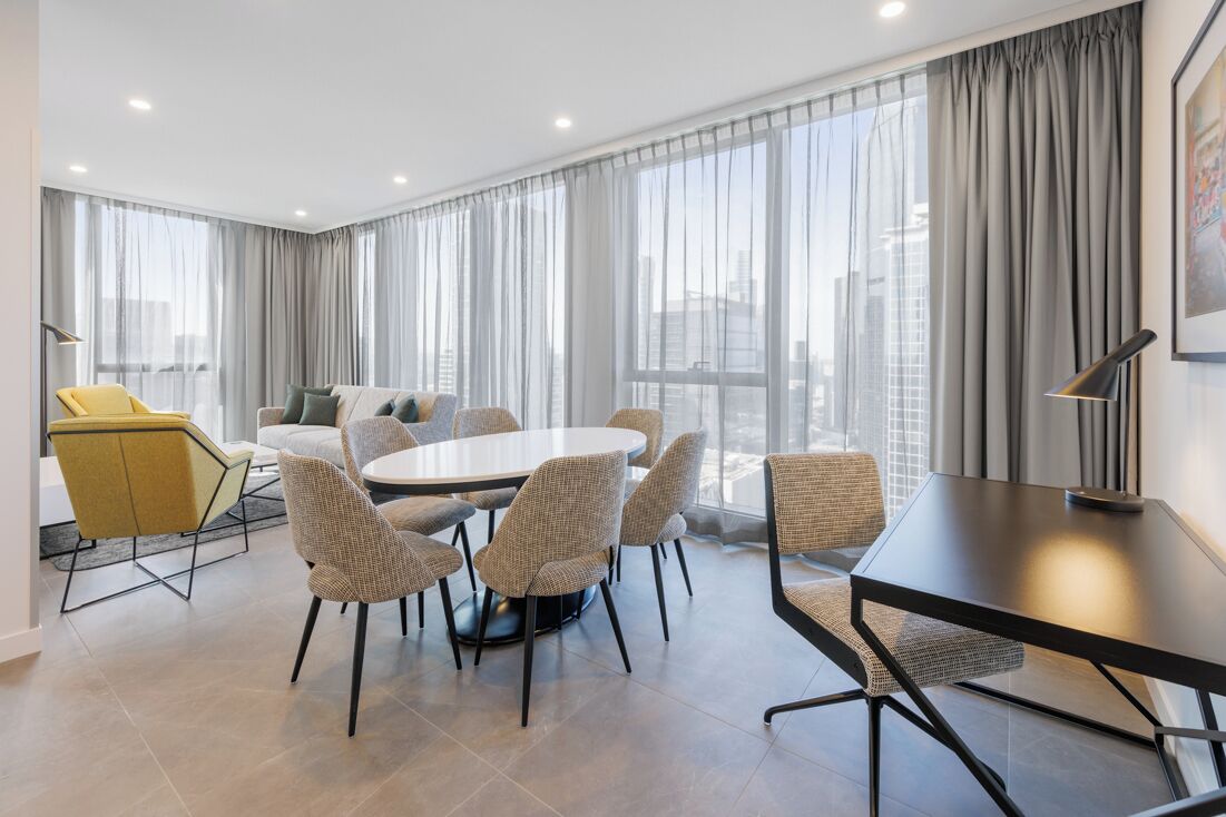 A modern hotel room with a round dining table, comfortable seating, large windows with curtains, and a city view.