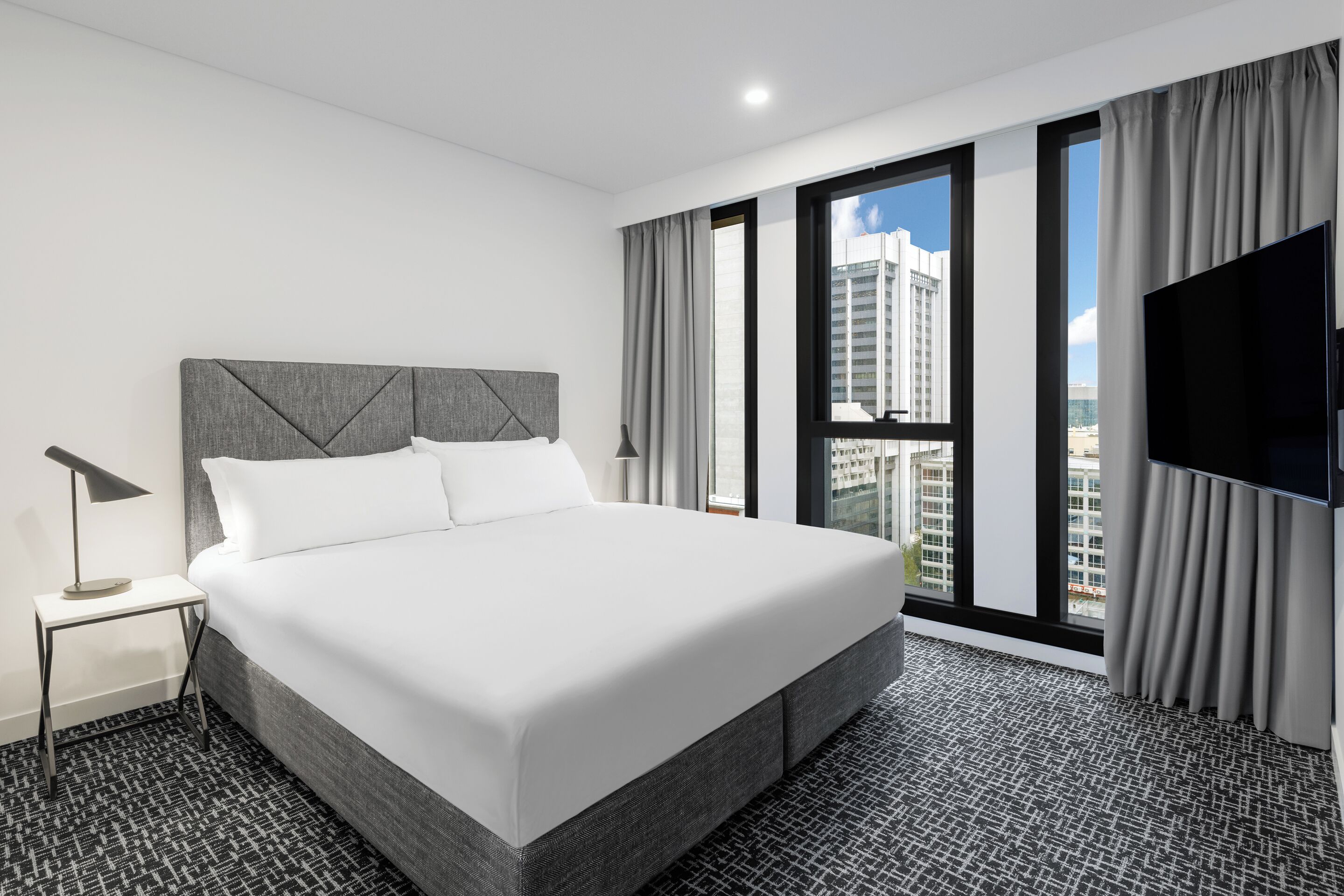 A modern hotel room with a large bed, a flat-screen TV, and a city view through large windows.