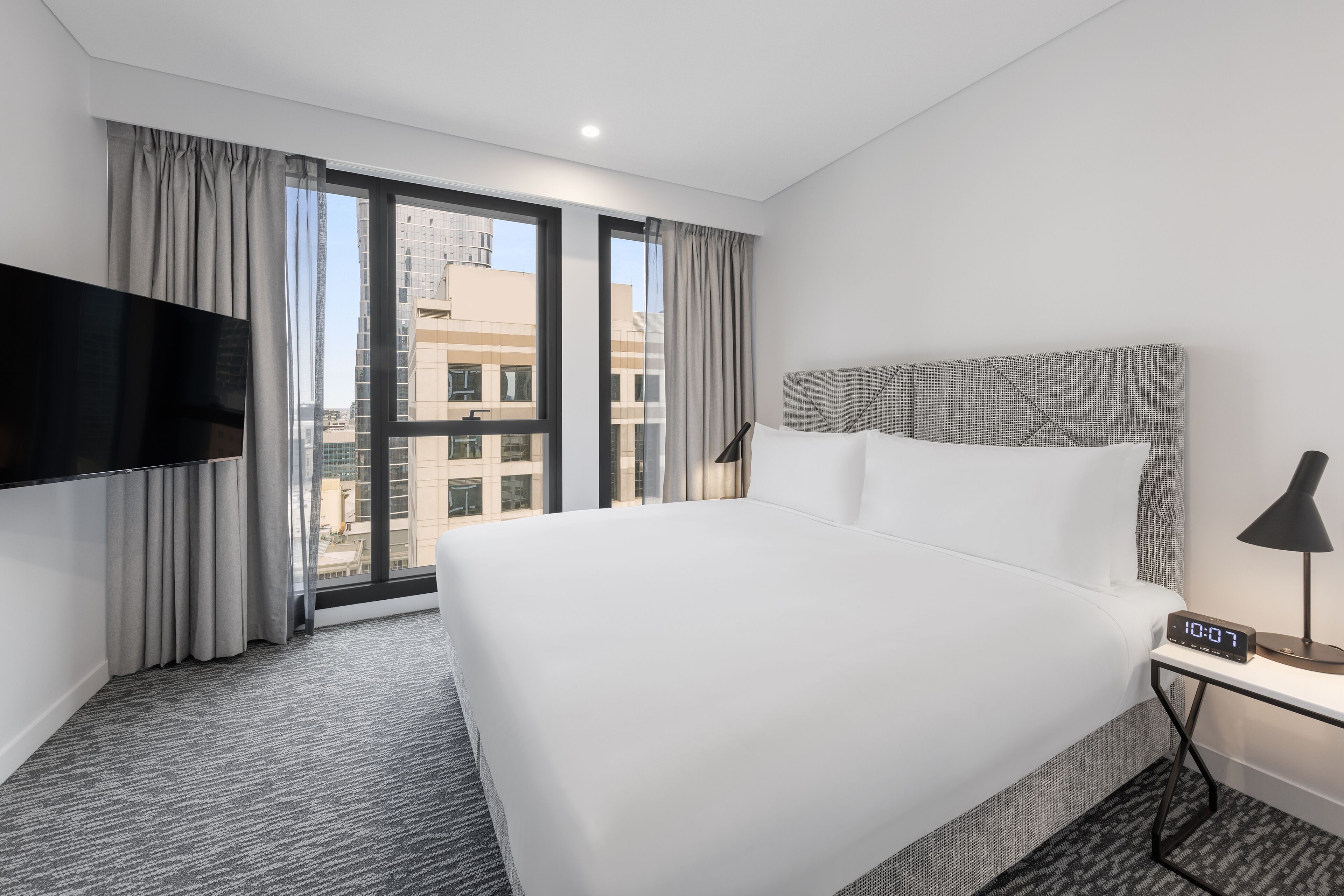 A modern hotel room with a large bed, a TV mounted on the wall, a bedside table with a lamp, and a view of the city through large windows.