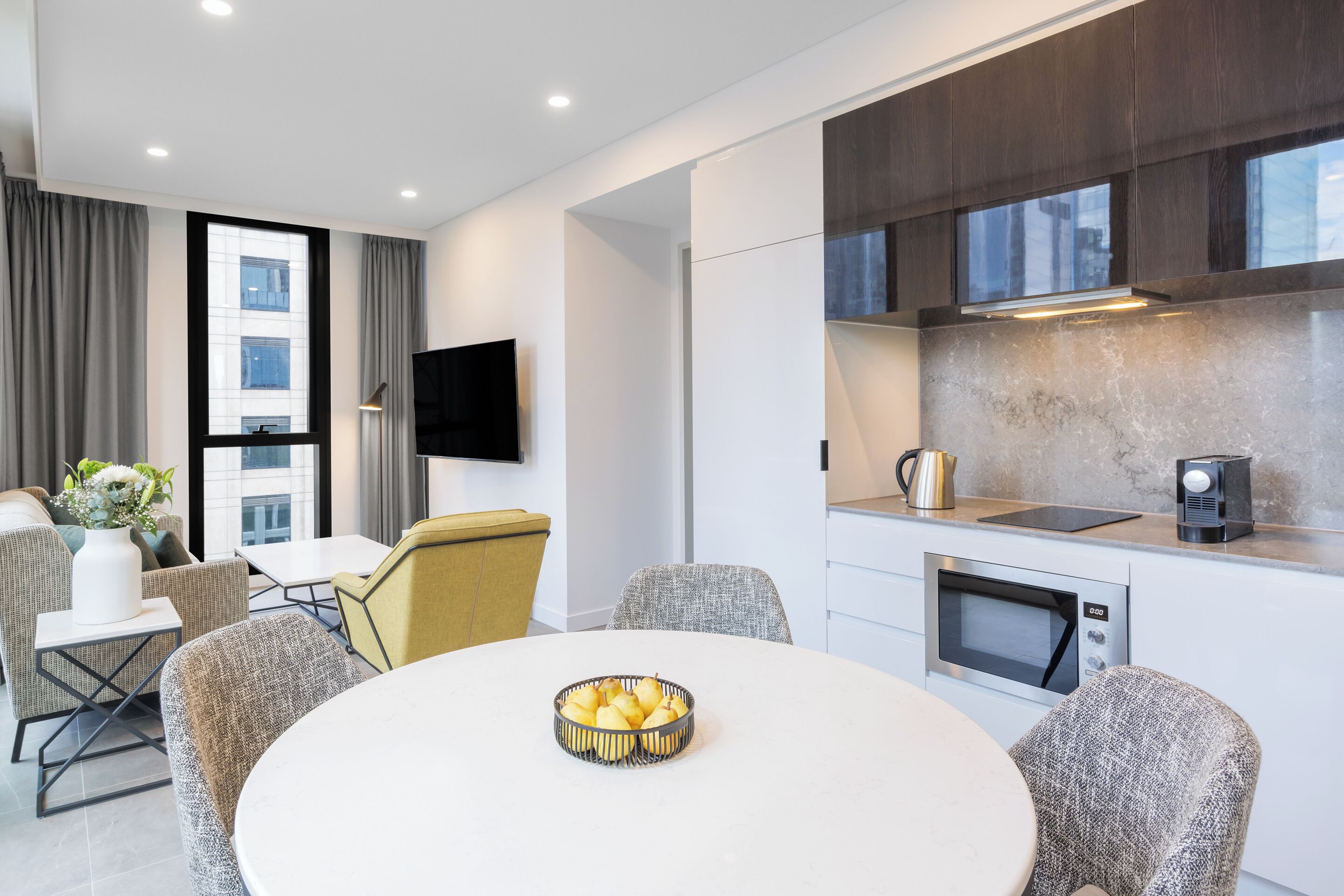 A modern hotel room with a dining area, a kitchen with a microwave, and a flat-screen TV.