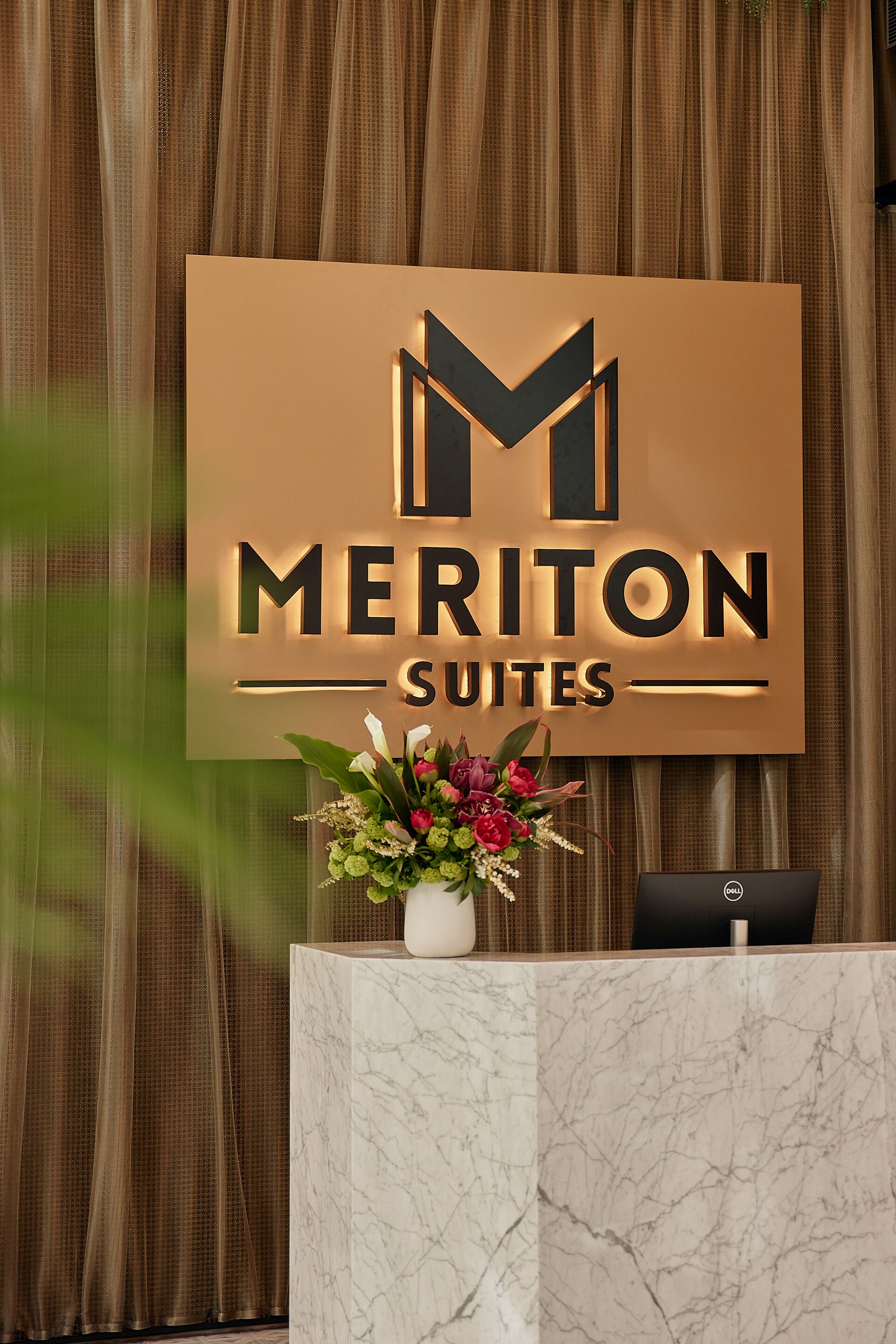 Un mostrador de recepción con un letrero de Meriton Suites, una planta en maceta y un ordenador Dell.