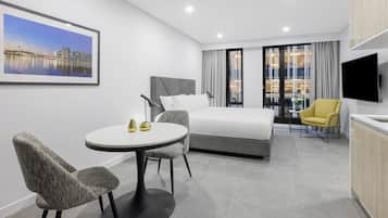 A modern hotel room with a bed, a dining table with chairs, a TV, and a large window with curtains.