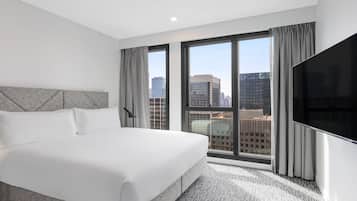 A hotel room with a large bed, a flat-screen TV, and a city view through large windows.