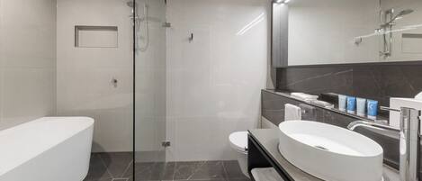 Un baño moderno con ducha de vidrio, bañera independiente y tocador con lavamanos grande y espejo.