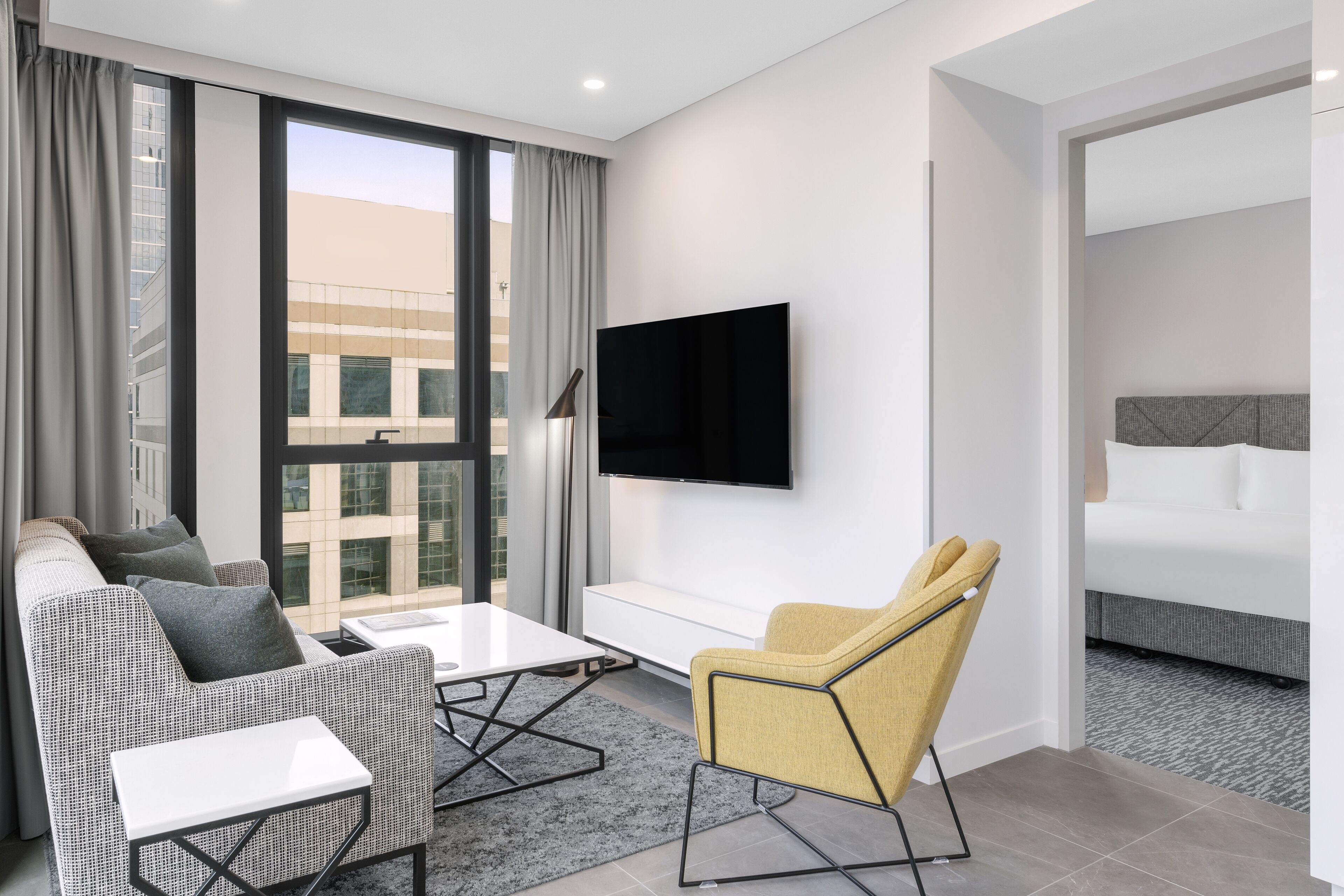 A modern hotel room with a sofa, a yellow armchair, a flat-screen TV, and a bed.
