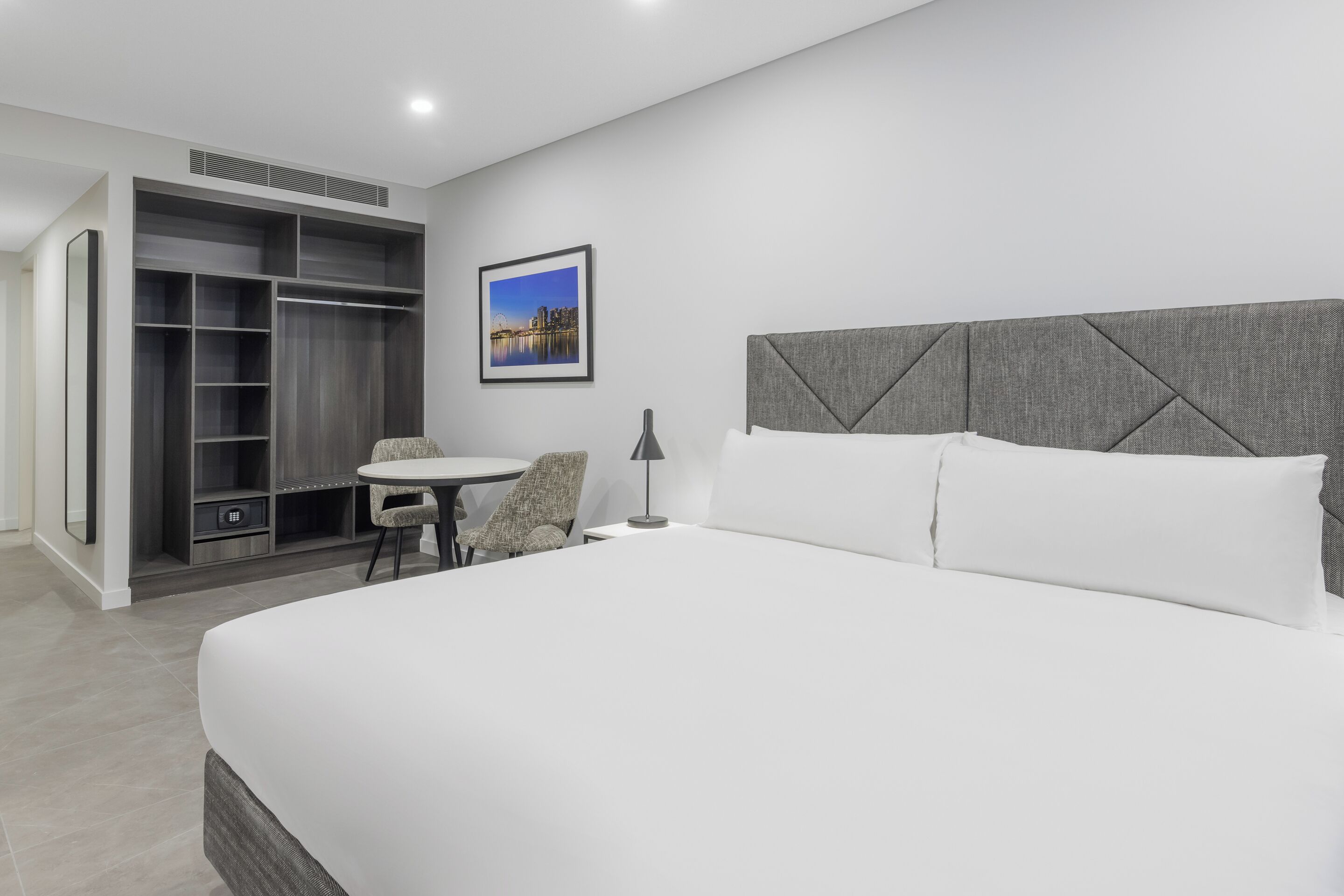 A modern hotel room with a large bed, a desk with a chair, a wardrobe, and a framed picture on the wall.