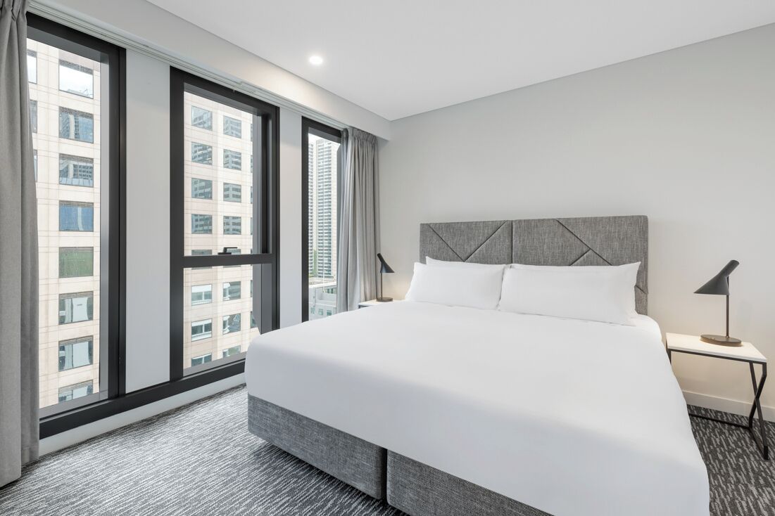 A hotel room with a large bed, a nightstand, a lamp, and a view of tall buildings through large windows.