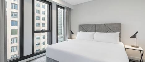 Une chambre d’hôtel avec un grand lit, une table de chevet, une lampe et une vue sur de hauts immeubles, visible à travers de grandes fenêtres.
