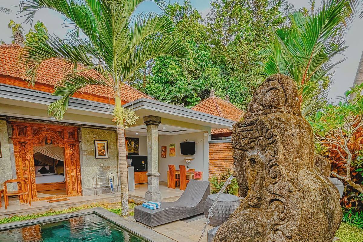 A pool area with a stone statue, a covered seating area, and a building with a tiled roof.
