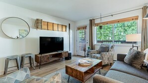 A modern living room with a flat-screen TV, wooden coffee table, patterned sofas, and a large window with a view of greenery.