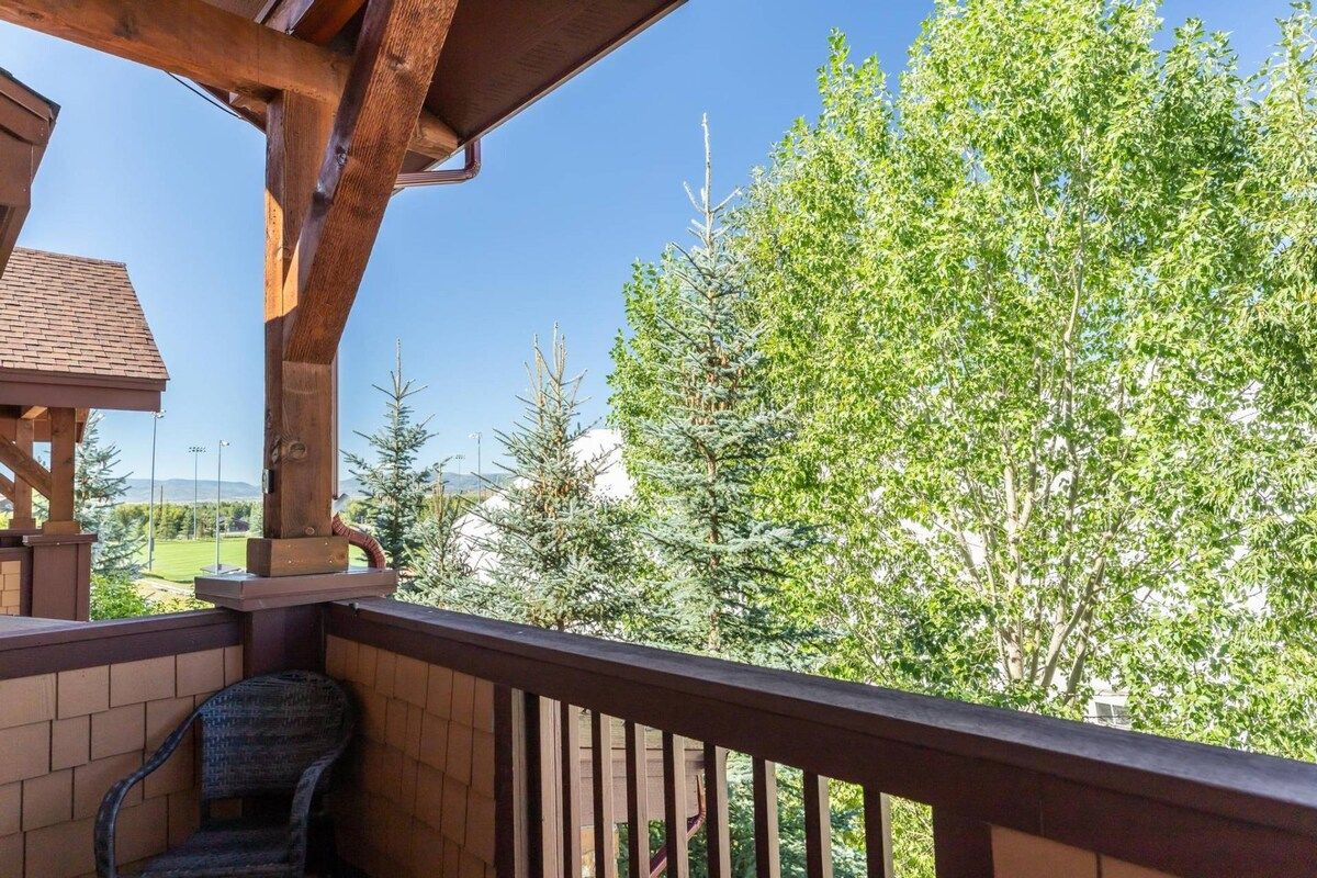 A balcony with a wooden railing, a chair, and a view of trees and a building in the background.