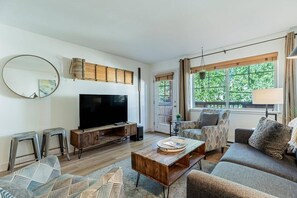 Una sala de estar con una televisión de pantalla plana, una mesa de centro de madera, sofás estampados y un ventanal con cortinas.