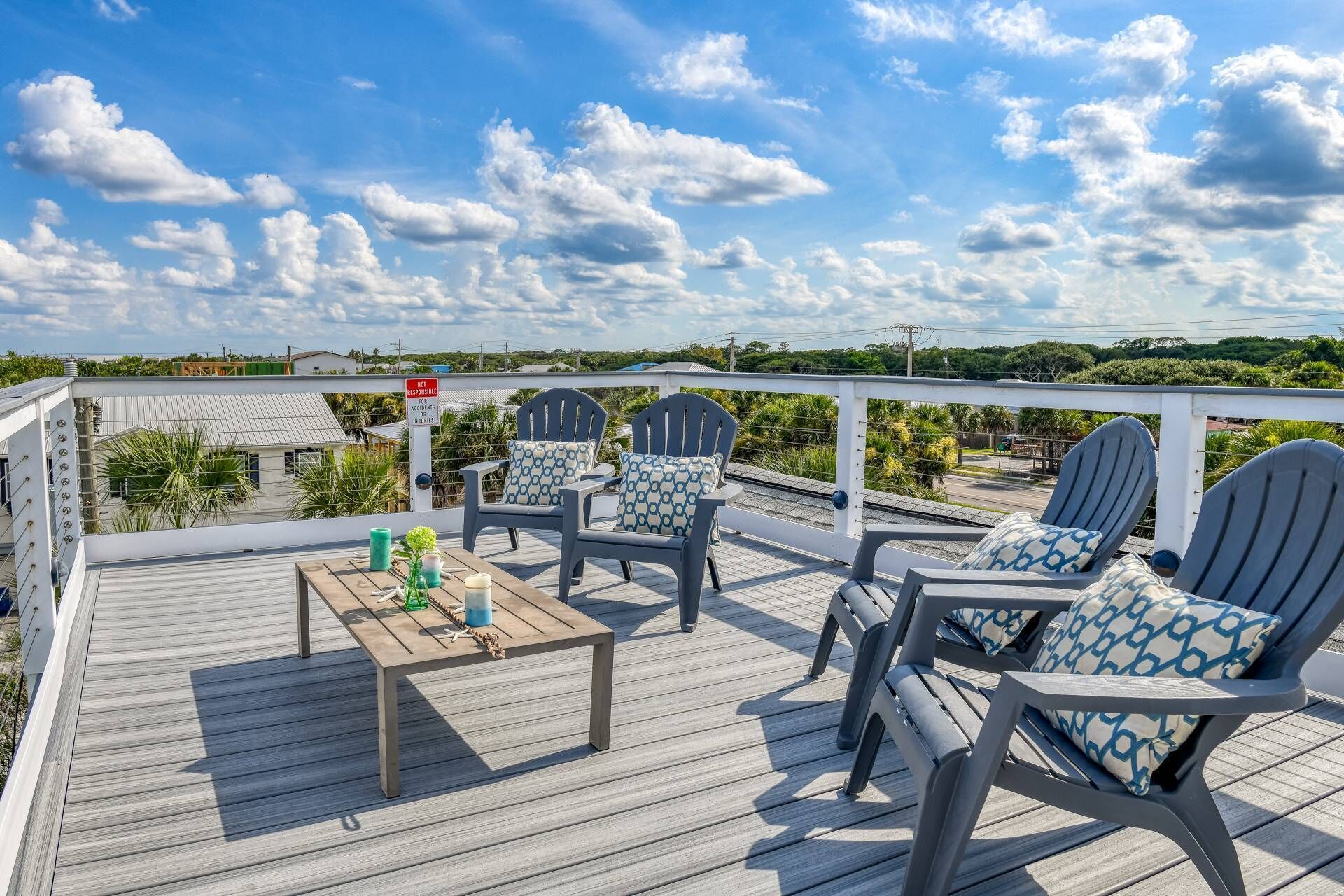 A wooden deck with two Adirondack chairs and a small table, overlooking a road and greenery.